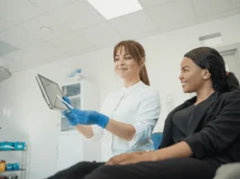 A smiling health care professional wearing blue gloves shows a patient medical information on a tablet in a clinical setting